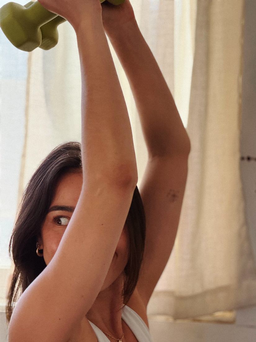 A woman lifting a green dumbbell overhead by a sunlit curtained window